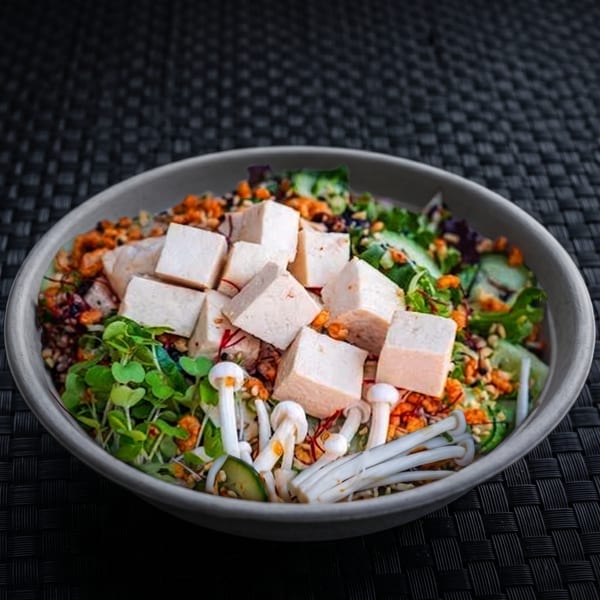 A vibrant salad bowl with tofu cubes, microgreens, mushrooms, and other toppings on a dark woven mat.